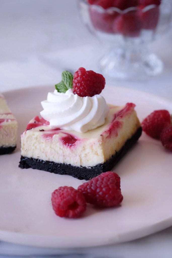Slice of white chocolate raspberry cheesecake bar topped with whipped cream, a mint leaf, and raspberry