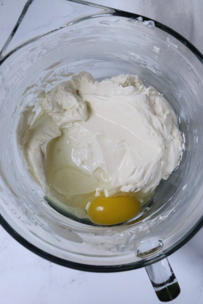 Cheesecake batter in a glass mixing bowl, with one whole egg