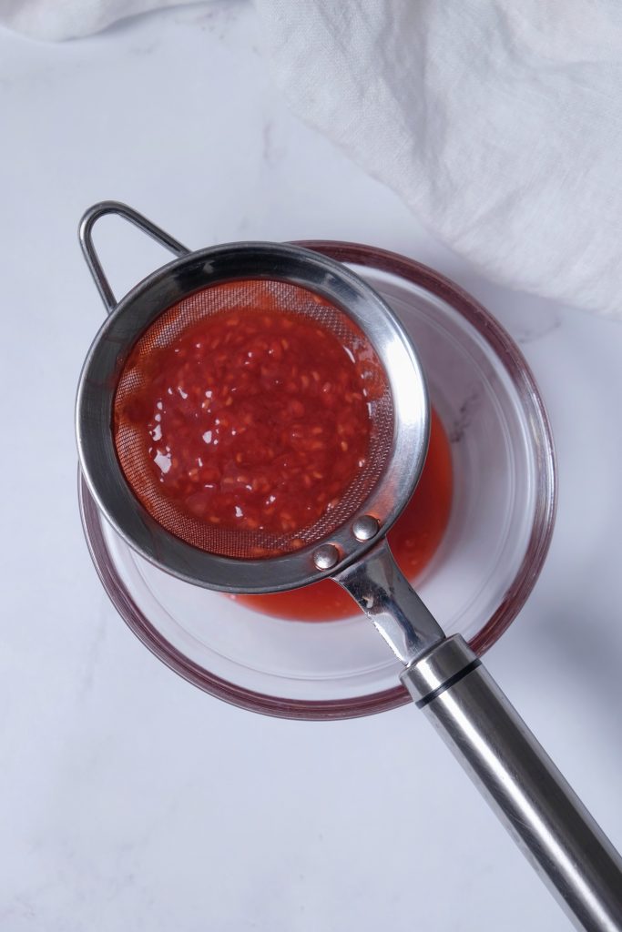Raspberry sauce in a mesh strainer over a glass bowl